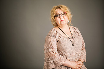 Kirjailija Niina Hakalahti istuu ruskehtavassa pitsimekossa ja katsoo kameraa. Hänellä on punertavat silmälasit, keltainen tukka ja riipus nahkaisessa hihnassa.