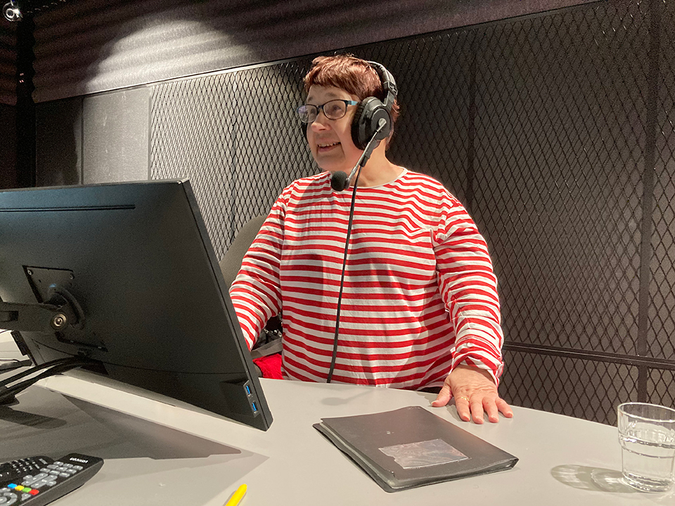 Rovasti Tuula Paasivirta punavalkoraitaisessa puserossaan kuulokkeet korvillaan näytön edessä pitämässä radiohartautta.